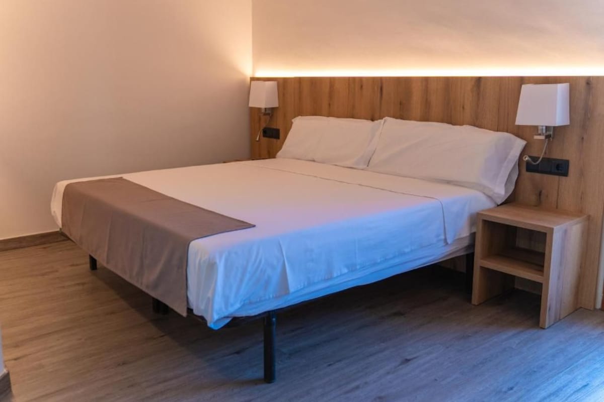 Habitación individual con cama de una sola plaza, cabecera de madera, mesitas de noche y lámparas fijadas a la pared.