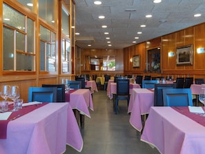 Interior de restaurante con varias mesas, sillas y vista a la calle a través de amplios ventanales.