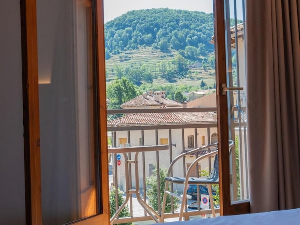 A room with a view of a hillside, a balcony with a chair, and a building with a tiled roof.