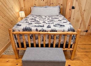 A wooden bed with a forest-themed bedspread and a matching headboard, a bedside lamp, and a bench at the foot of the bed. - The Reel Escape (Dahlonega)