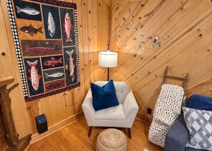A cozy room with a wooden ceiling, a white armchair with a blue pillow, a patterned armchair, a wall-mounted lamp, and a fish-themed artwork. - The Reel Escape (Dahlonega)