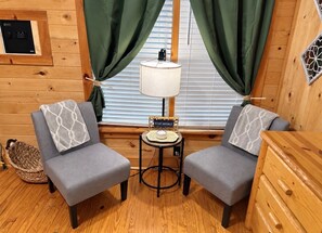 A room with two grey armchairs, a small round table with a lamp, a wooden cabinet, and a basket on the floor. - The Reel Escape (Dahlonega)
