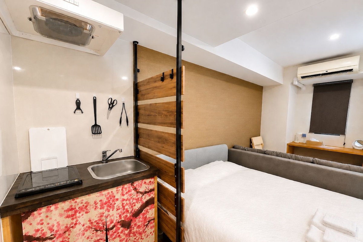 Quarto de hotel compacto, com cama, pequena cozinha, pia e janela com unidade de ar condicionado.