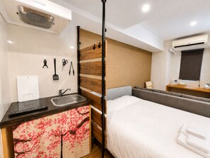 Quarto de hotel compacto, com cama, pequena cozinha, pia e janela com unidade de ar condicionado.