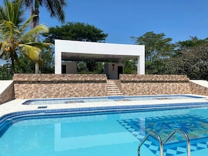 En pool med ett geometriskt mönster av kakelplattor, en vit byggnad med en täckt uteplats och en stenmur.