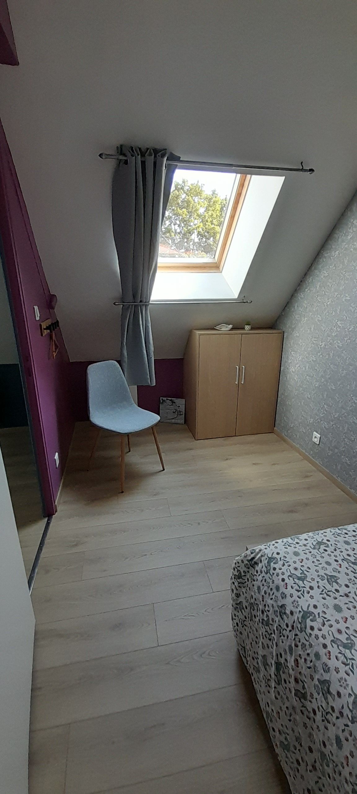 A room with a sloped ceiling, a skylight, a chair, a wardrobe, and a bed with a patterned cover.
