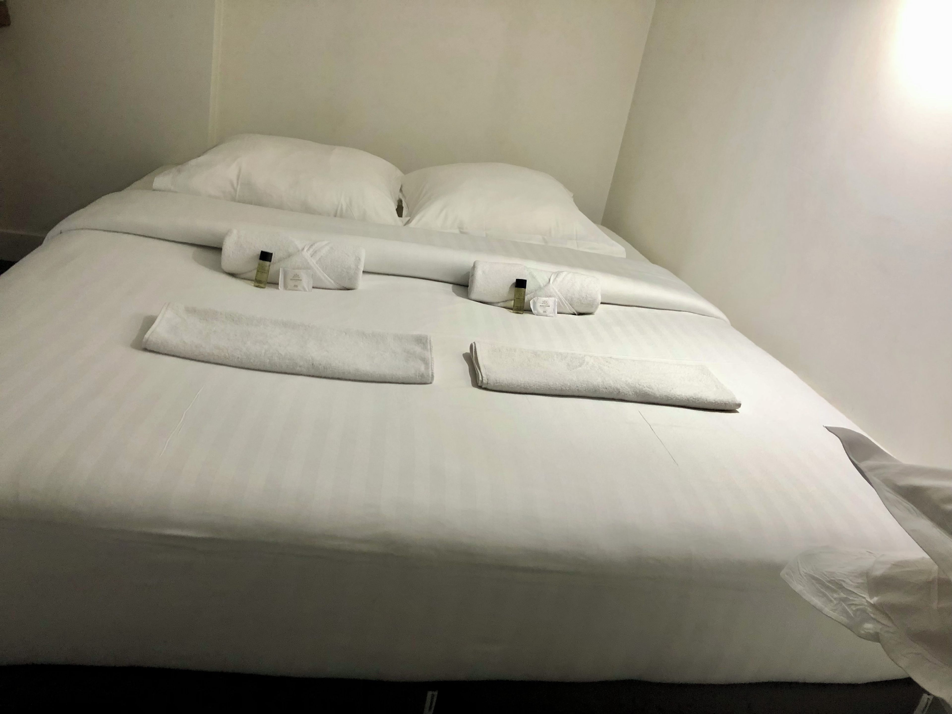 A neatly made bed with white linens and towels, a bottle of lotion, and a small toiletry bag.