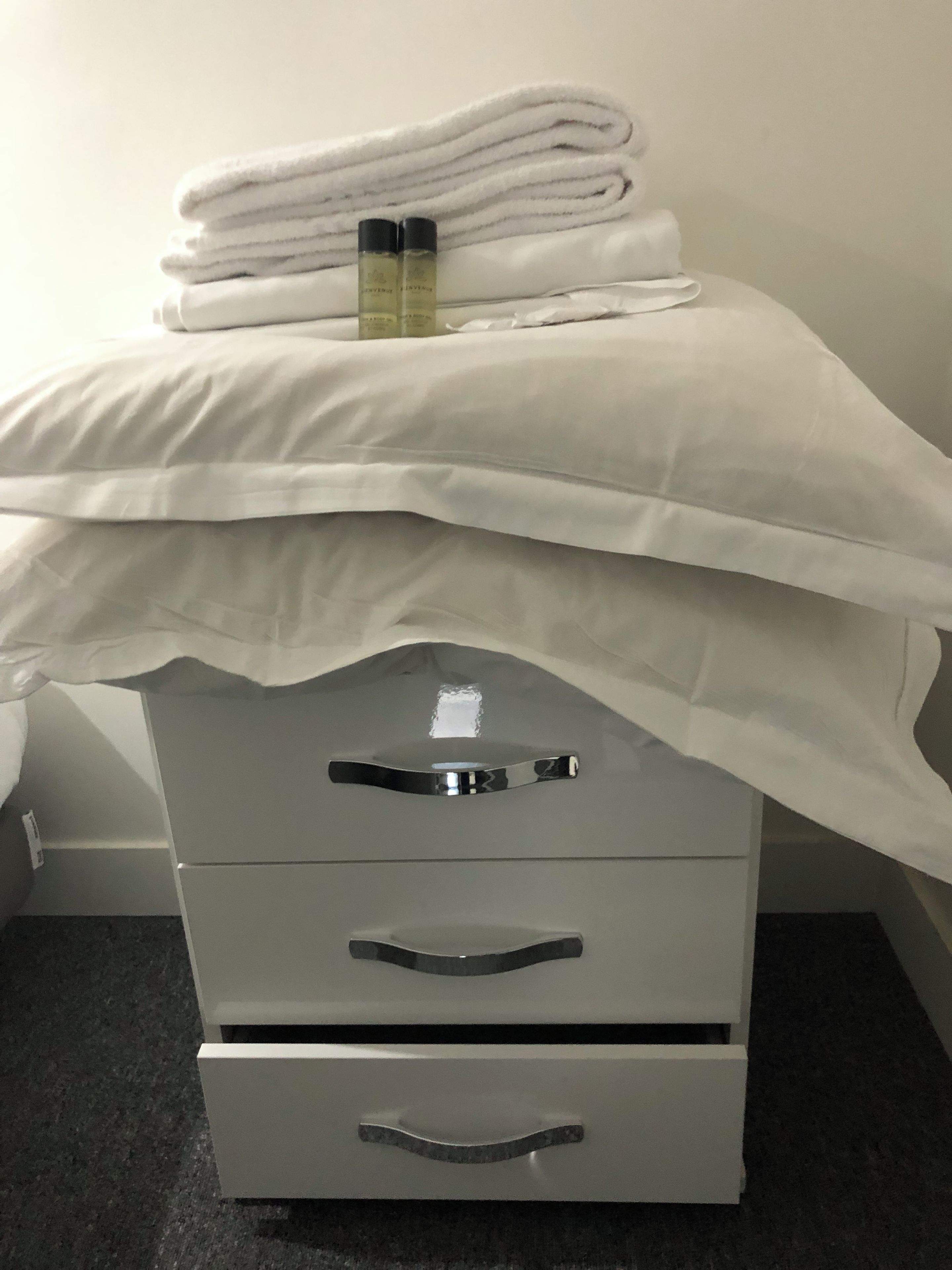 A bed with white linens and two bottles on top, a grey dresser with drawers, and a plain wall in the background.