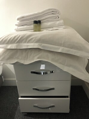 A bed with white linens and two bottles on top, a grey dresser with drawers, and a plain wall in the background. - Vrbo Property (Paris)