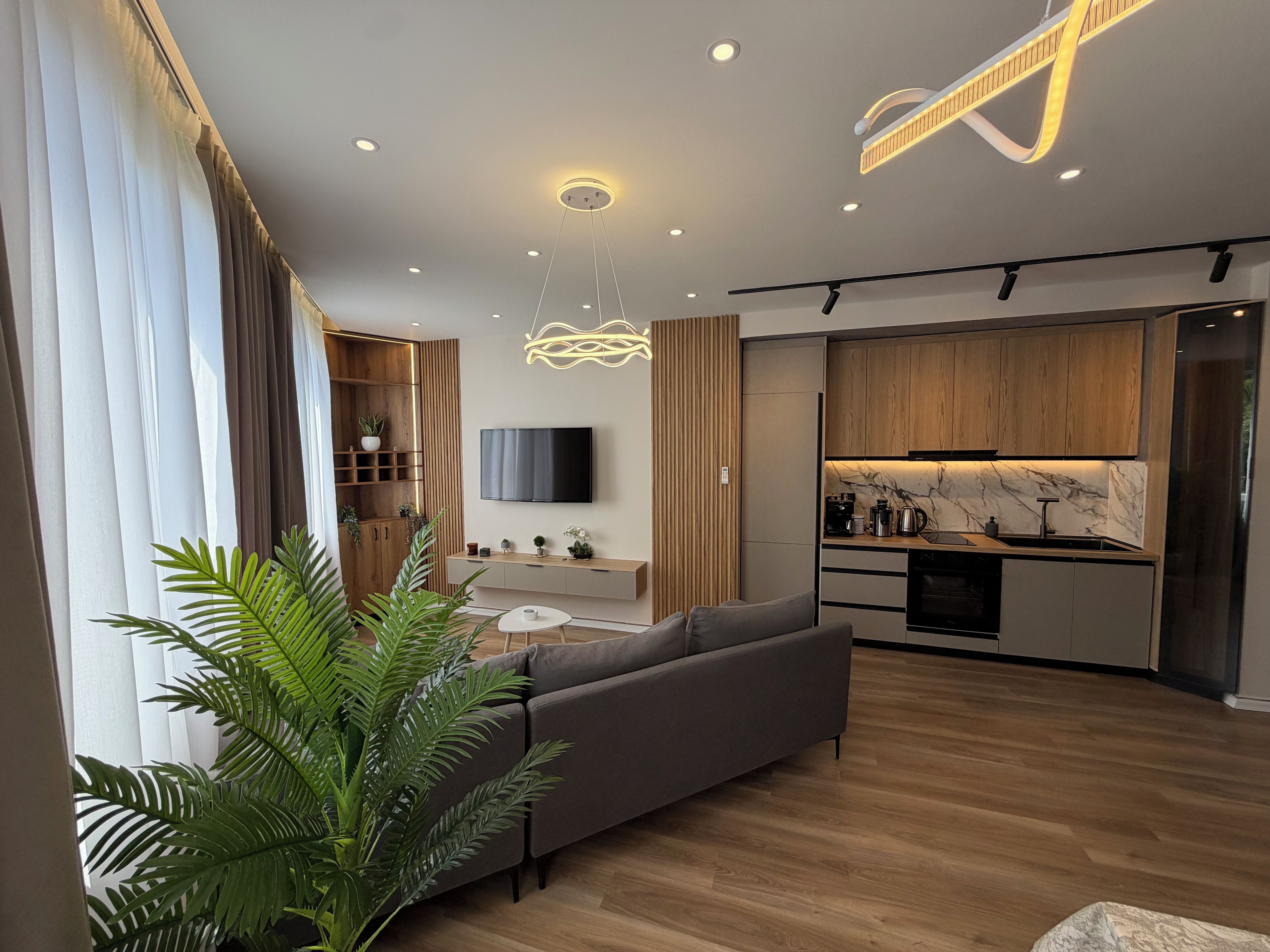 Ein modernes Wohnzimmer mit einem grauen Sofa, einem Flachbildfernseher an der Wand und einer Holz-Küche mit Marmor-Spritzschutz.