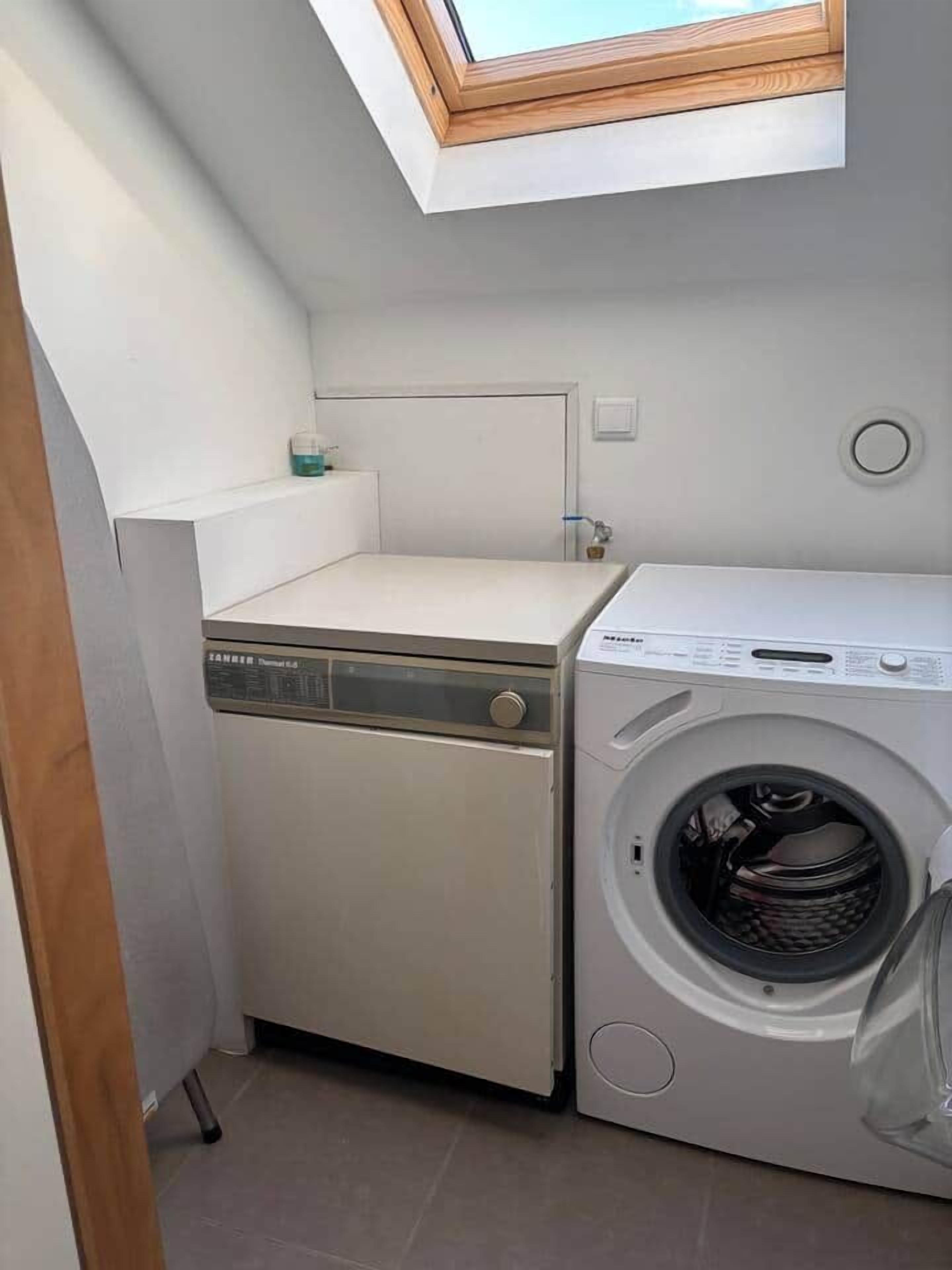 A compact laundry room with a washing machine and a dryer, a skylight above, and a small shelf with a container.