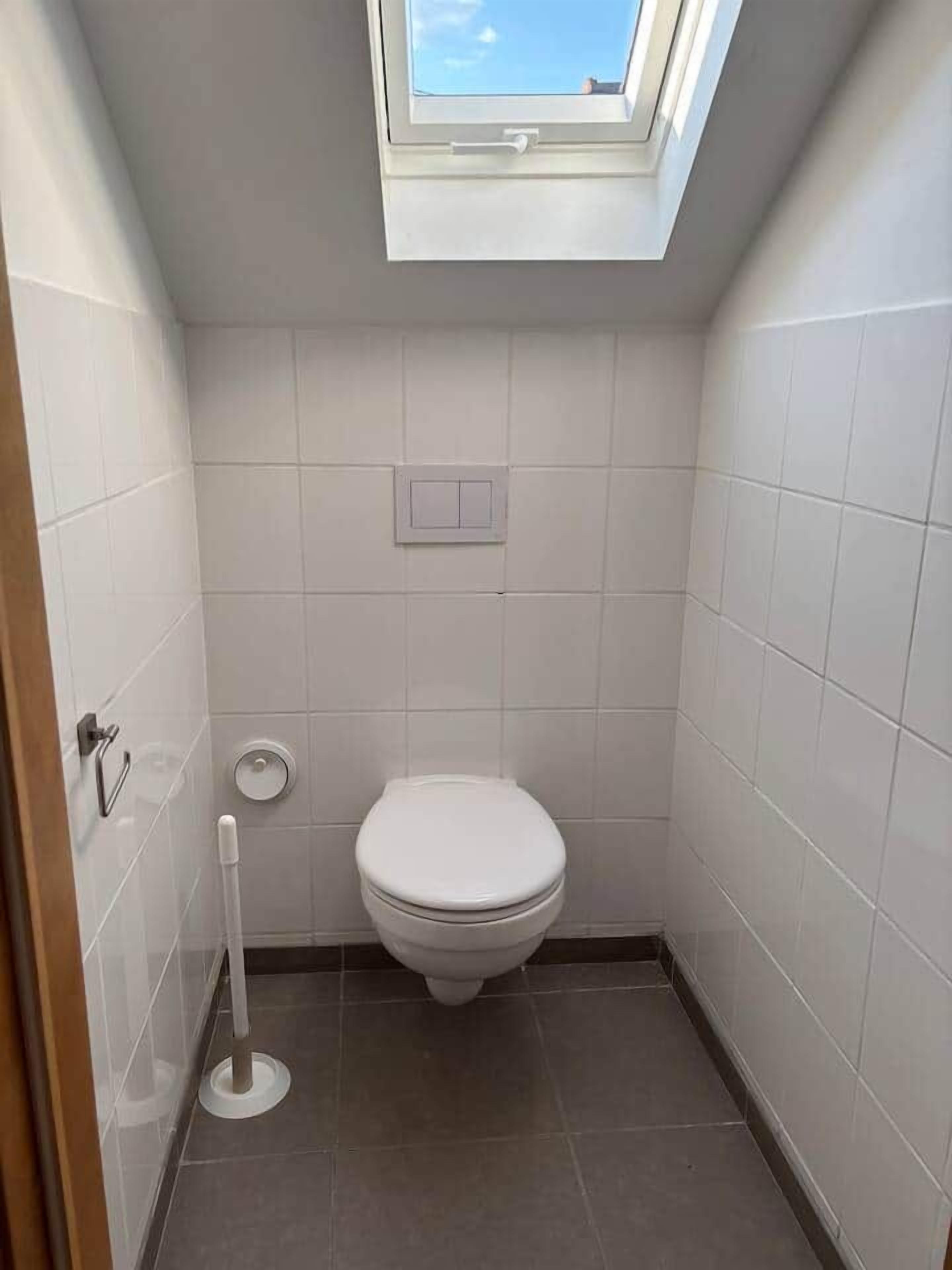 Un baño pequeño con azulejos blancos y una claraboya sobre el inodoro.