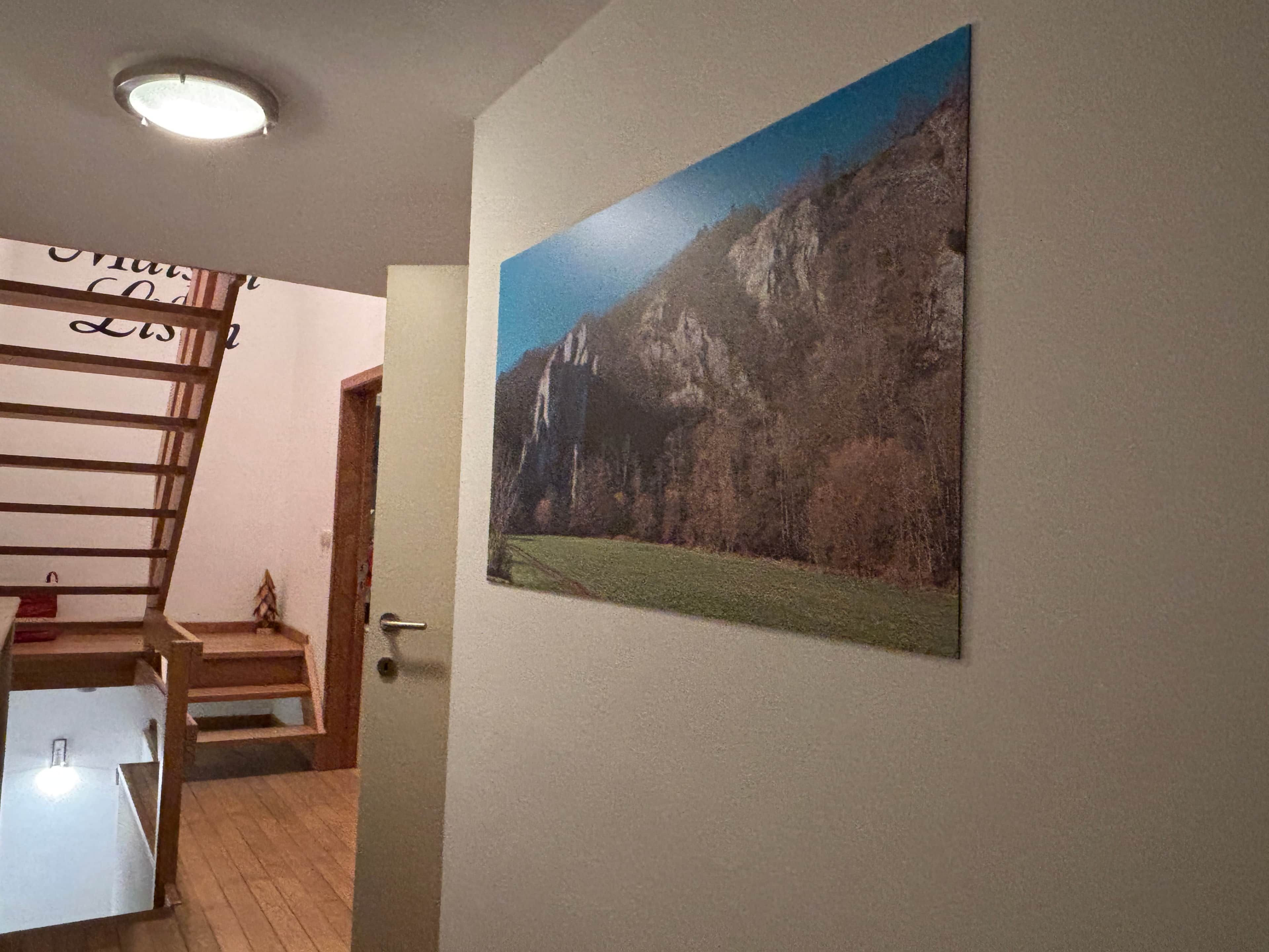 Een trap die leidt naar een kamer met een deur, een aan de muur hangend schilderij van een berglandschap en een plafondlamp.
