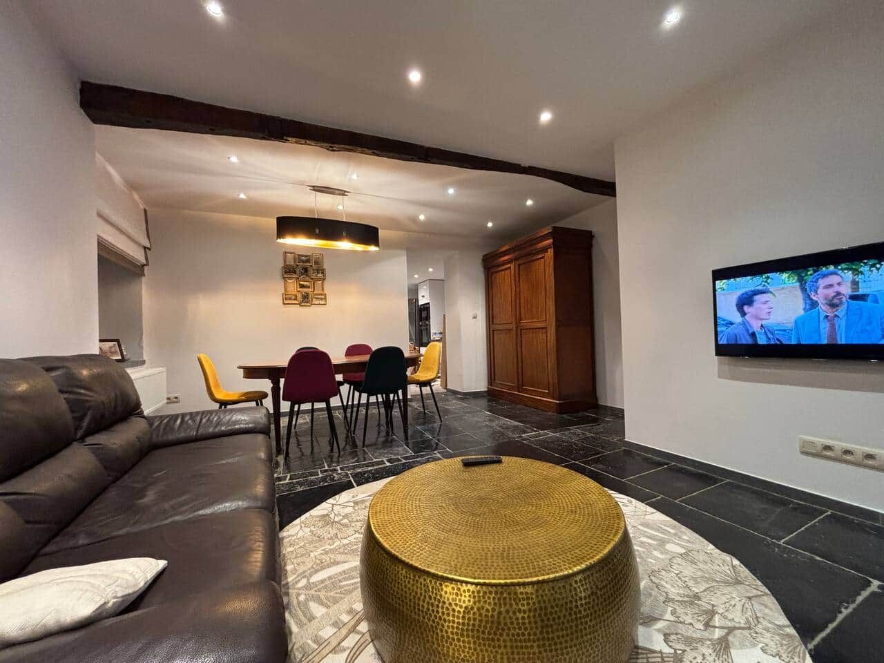 A modern living room with a sofa, a round coffee table, a dining area with chairs, and a flat-screen TV mounted on the wall.