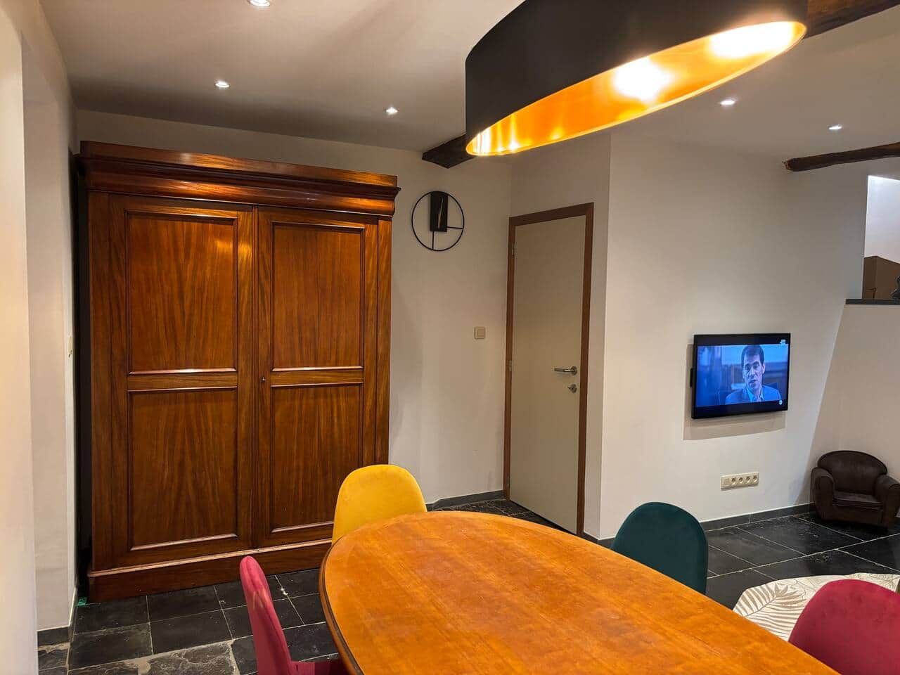Een kamer met een houten kast, een aan de muur bevestigde televisie en een eettafel met stoelen.