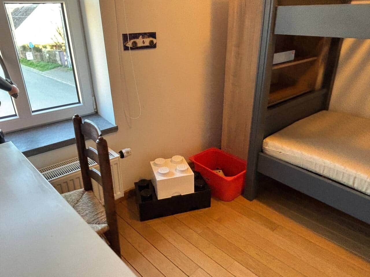 Una cama litera con colchón, una silla de madera, una mesita pequeña, un radiador, una ventana y una cesta roja.
