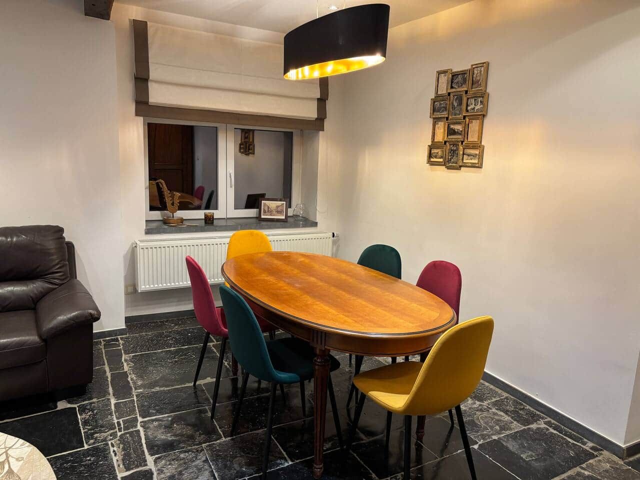 Un coin repas avec une table en bois et des chaises colorées, un canapé en cuir noir et un radiateur en arrière-plan.