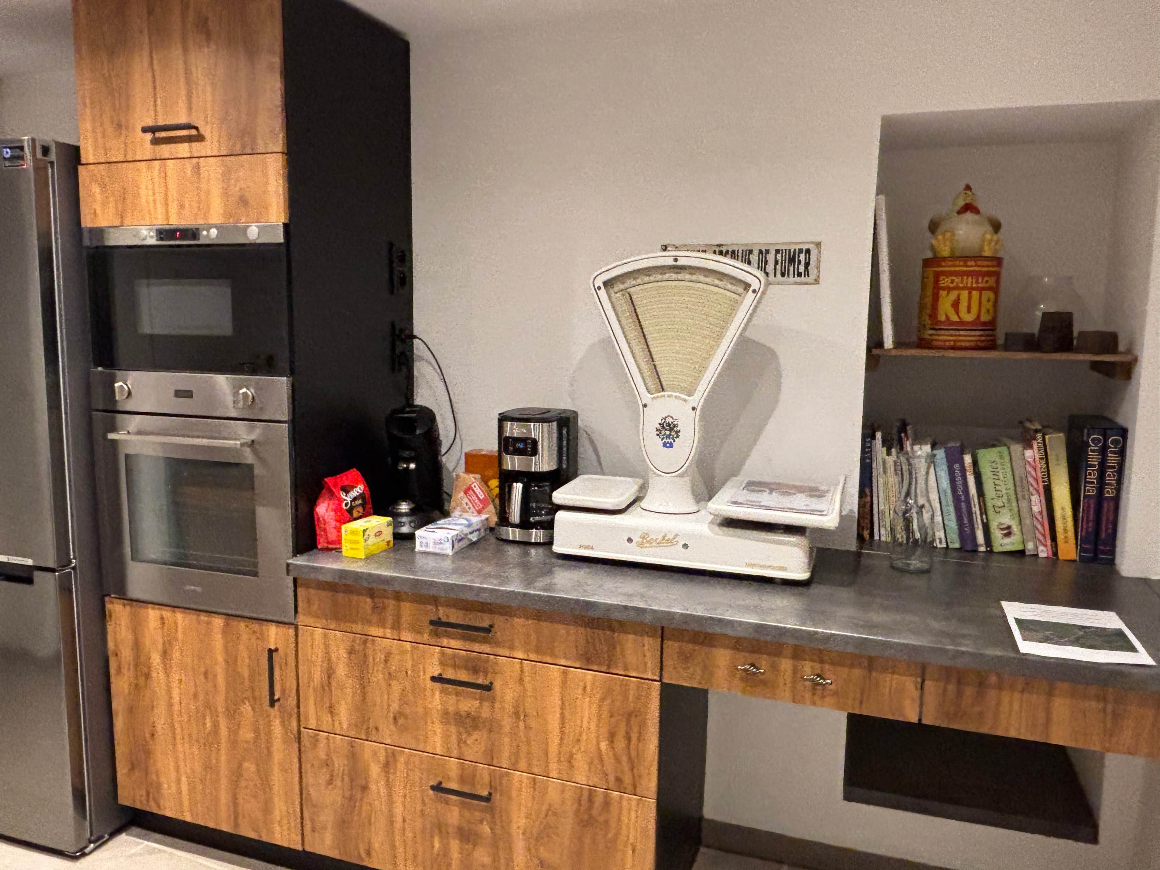 Een keuken met houten kastjes, een roestvrijstalen oven, een koffiezetapparaat, een weegschaal, en een plank met boeken en een voorraadbus.