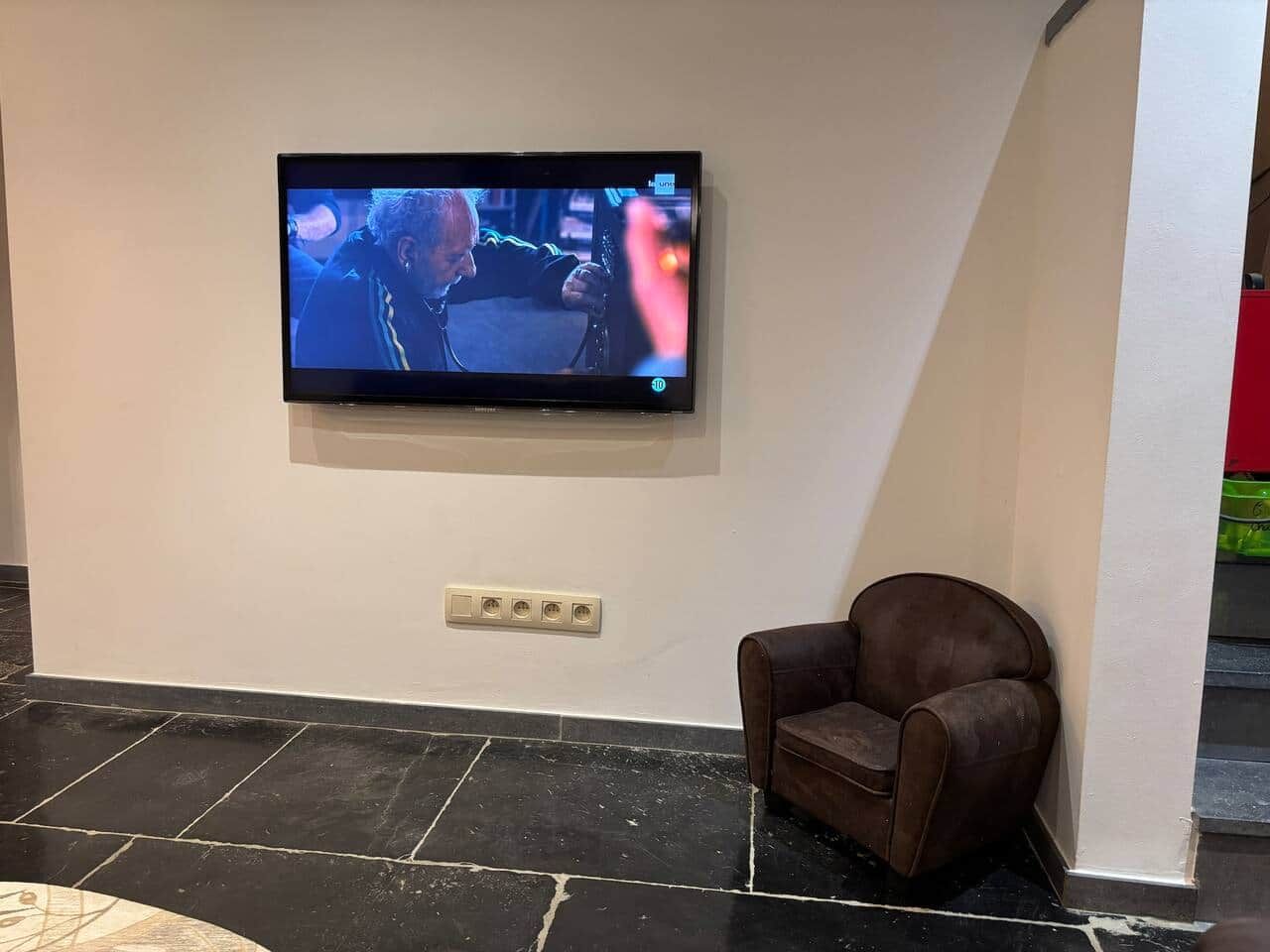 Een televisie hangt aan de muur, voor een fauteuil van bruin leer.