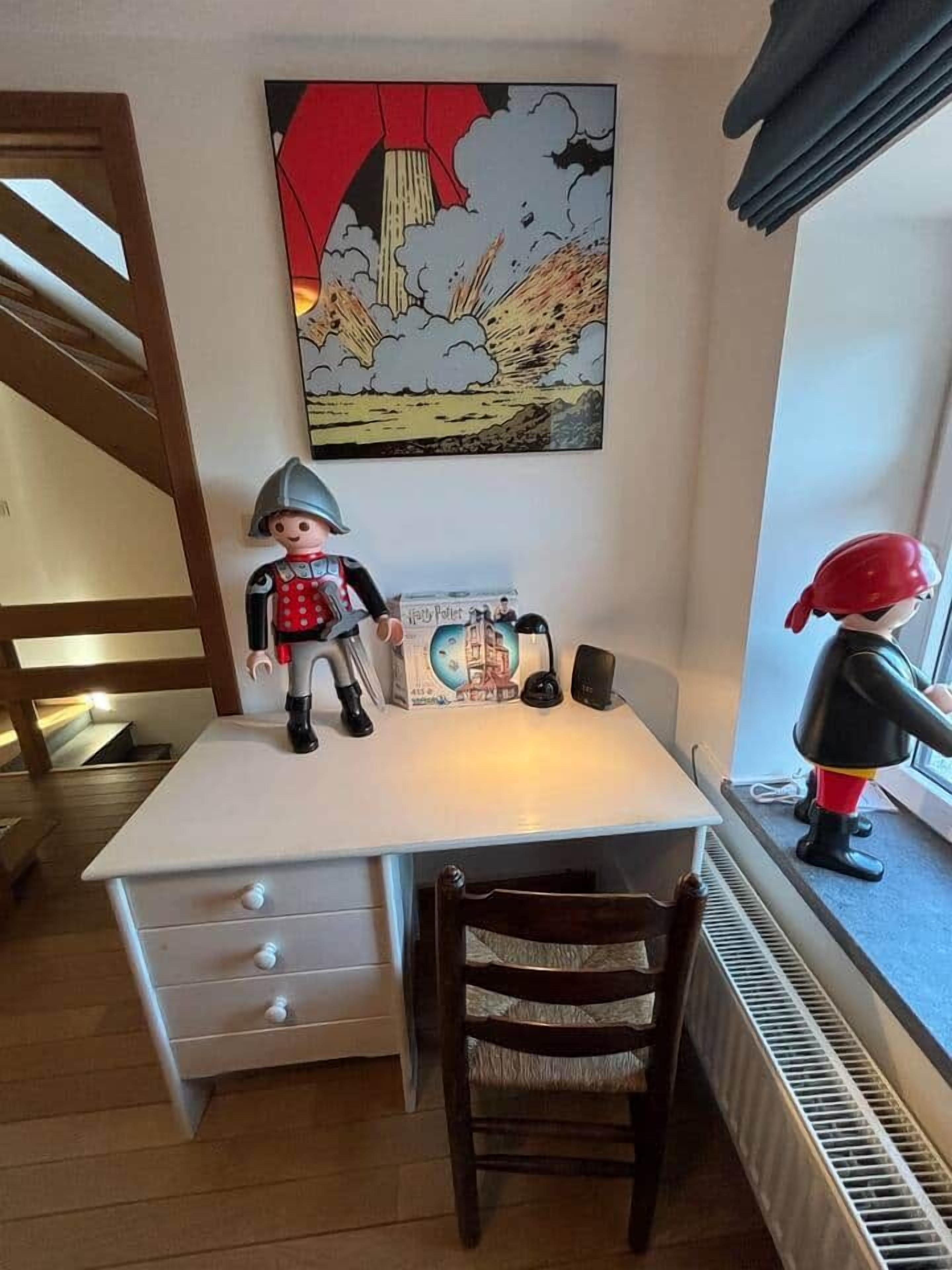 Une chambre d’enfant avec un bureau, deux figurines, une bande dessinée et une fenêtre.