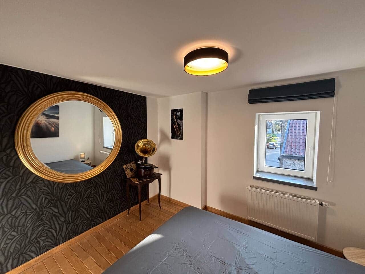 Une chambre à coucher avec un lit, un miroir rond, une petite table sur laquelle se trouve un gramophone, une fenêtre donnant sur un paysage agréable et un radiateur.