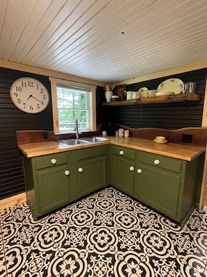 A kitchen with green cabinets, a wooden countertop, a black and white patterned floor, a wall clock, and a window.