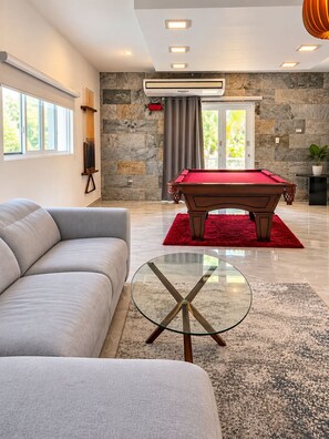 A modern living room with a pool table, a sofa, a glass coffee table, and a stone wall.