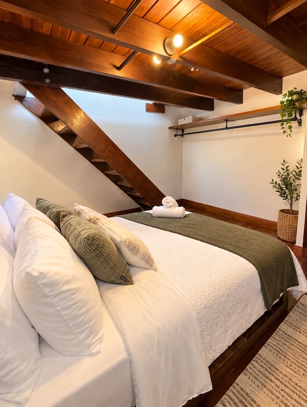 A bedroom with a sloped ceiling, a bed with white bedding, a green throw blanket, and a hanging plant.