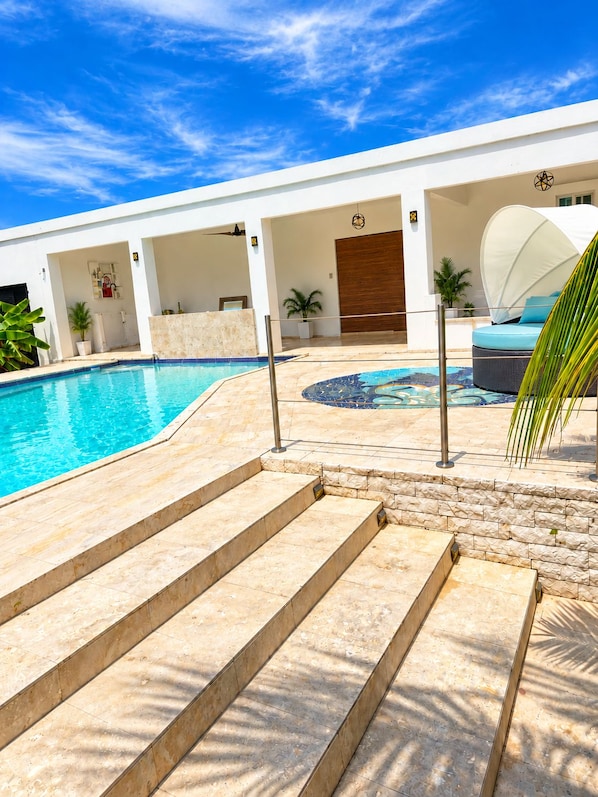 A swimming pool with steps leading to a covered seating area, surrounded by a white building with a wooden door.