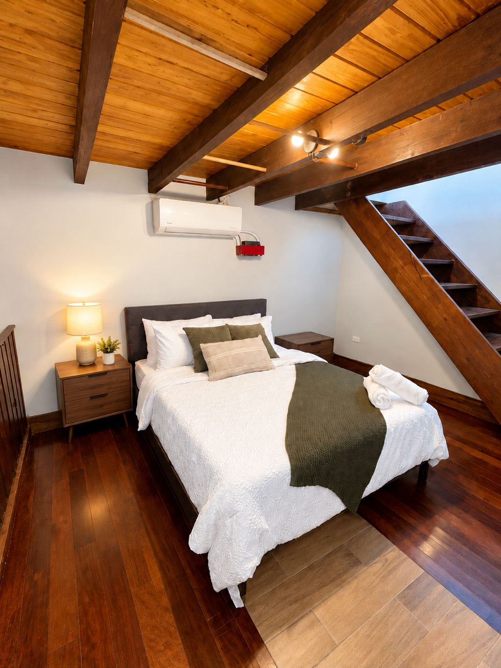 A bedroom with a wooden ceiling, a bed with white bedding, a nightstand with a lamp, and a staircase.