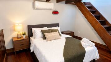 A bedroom with a wooden ceiling, a bed with white bedding, a nightstand with a lamp, and a staircase.