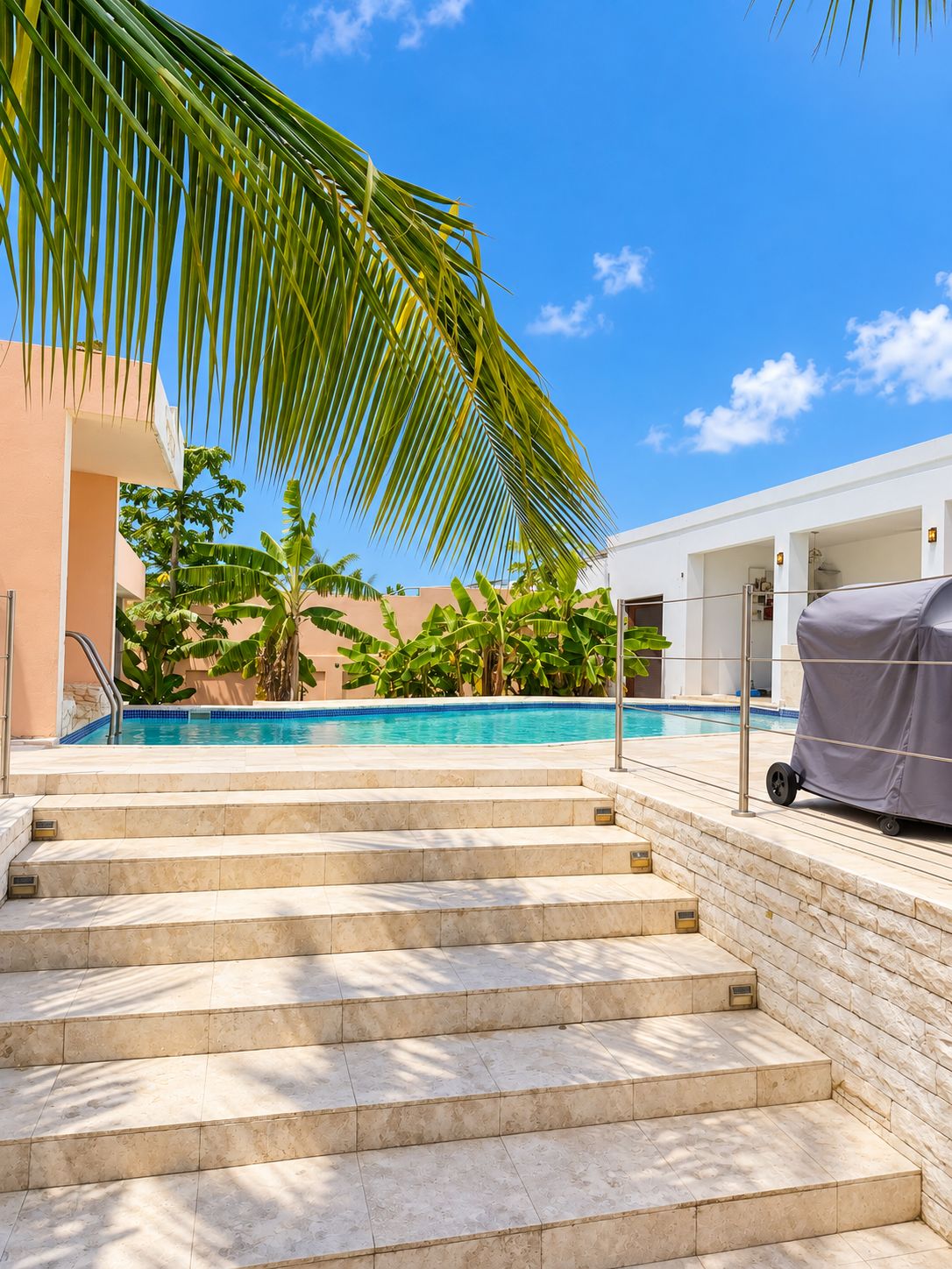 A swimming pool with steps leading down to the water, surrounded by palm trees and a building.