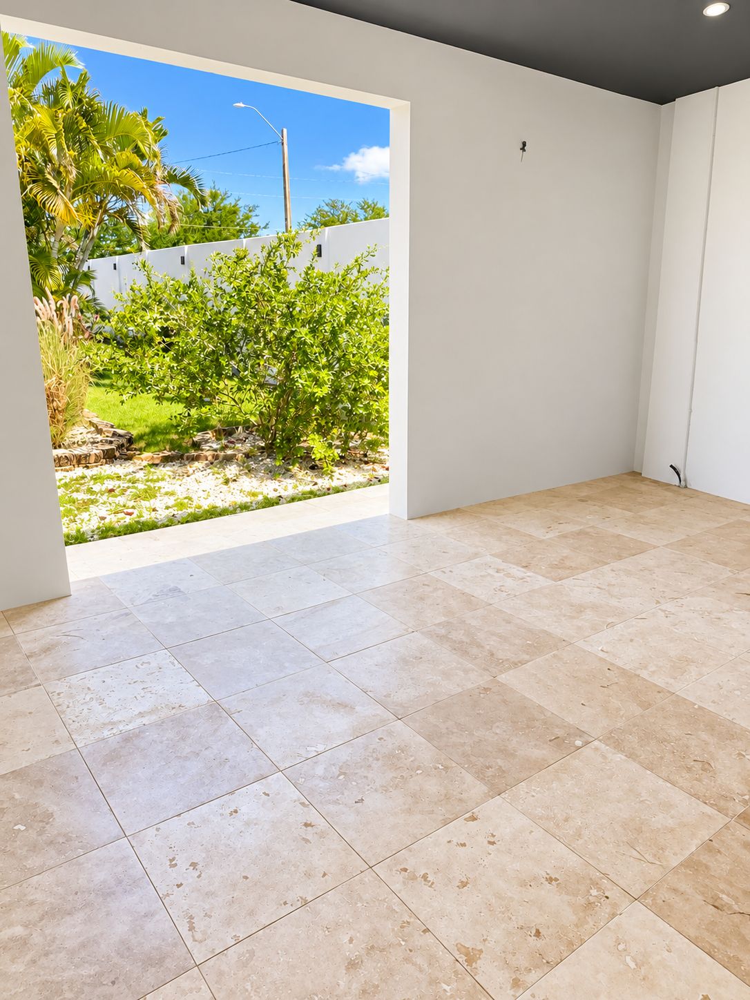 A room with a tiled floor and a view of a garden with palm trees and a white wall.