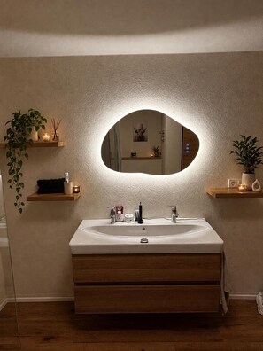 A bathroom with a large oval mirror, a white sink, wooden cabinets, and two wall-mounted shelves with plants and candles. - Cozy Vacation Rental Near Nuremberg (Petersaurach)