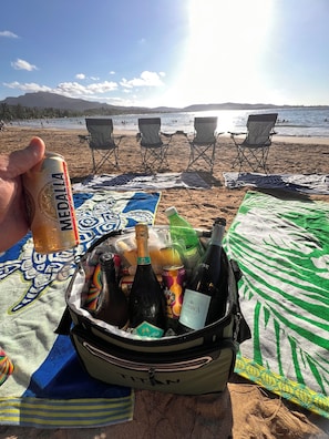 Eine Strandansicht mit einer Tasche, die Getränke und Snacks enthält, einer Bierdose und einer Weinflasche, mit Strandstühlen und dem Meer im Hintergrund.