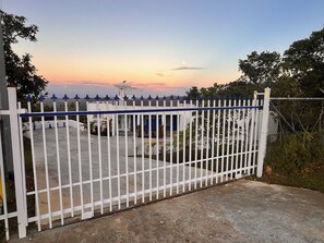 Un área cercada con valla blanca, un camino de hormigón y un edificio con techo azul al fondo.