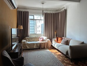 A living room with a sofa, a television, and a large window with curtains. - Paulista | Entire apartment of 170 m², 3 bedrooms and parking space (São Paulo)