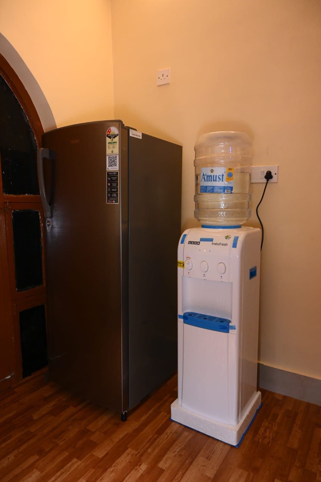 A refrigerator and a water dispenser in a room with wooden flooring and a door.