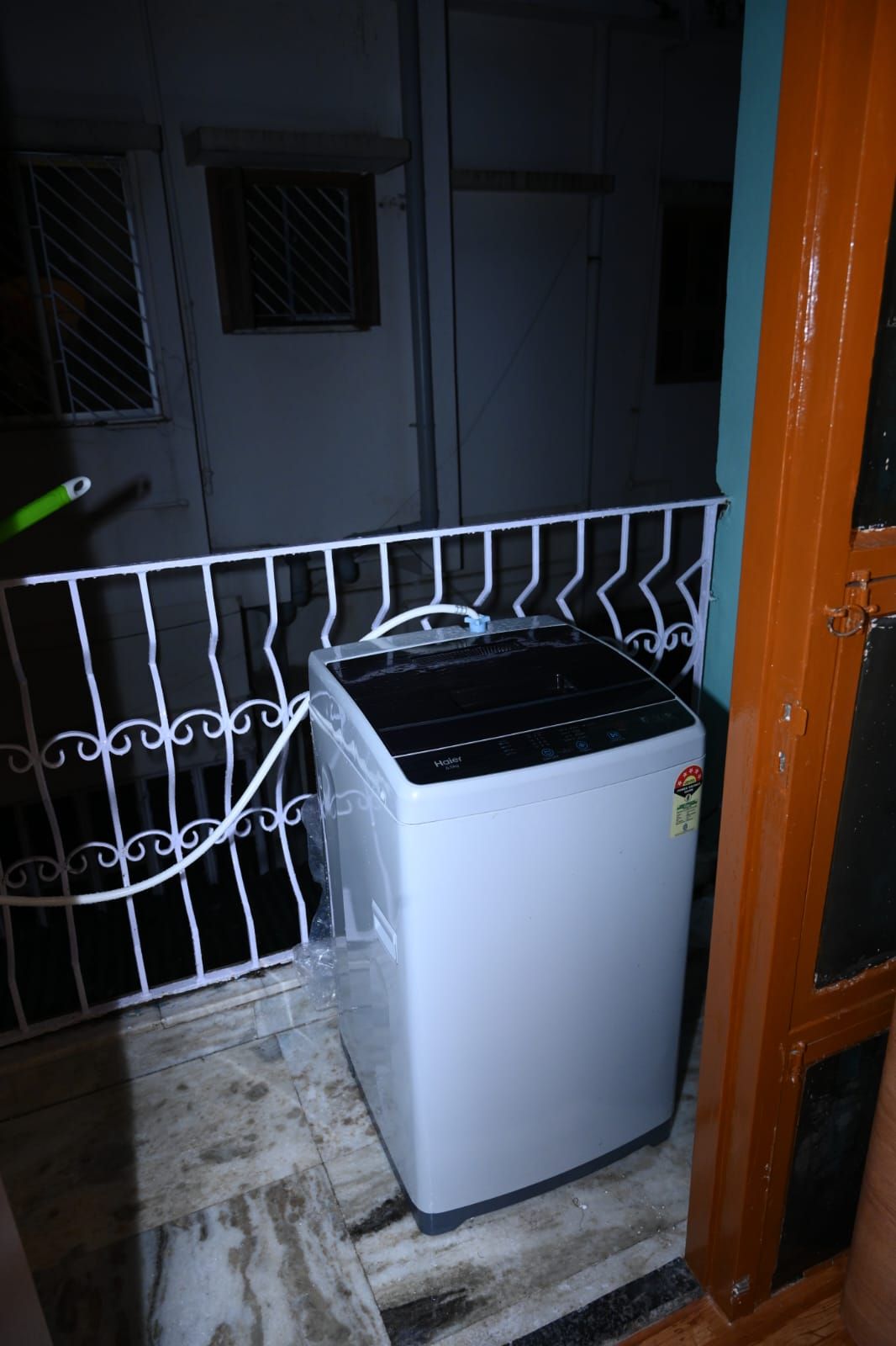 Ein kompaktes, weißes Gerät, vielleicht ein Kühlschrank oder eine Waschmaschine, steht in einer Ecke neben einer Metallreling und einer Tür mit Griff.