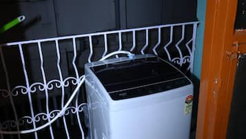Ein kompaktes, weißes Gerät, vielleicht ein Kühlschrank oder eine Waschmaschine, steht in einer Ecke neben einer Metallreling und einer Tür mit Griff.