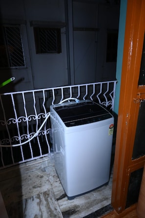 Un electrodoméstico pequeño y blanco, quizás un refrigerador o lavadora, ubicado en una esquina con baranda metálica y una puerta con manija.