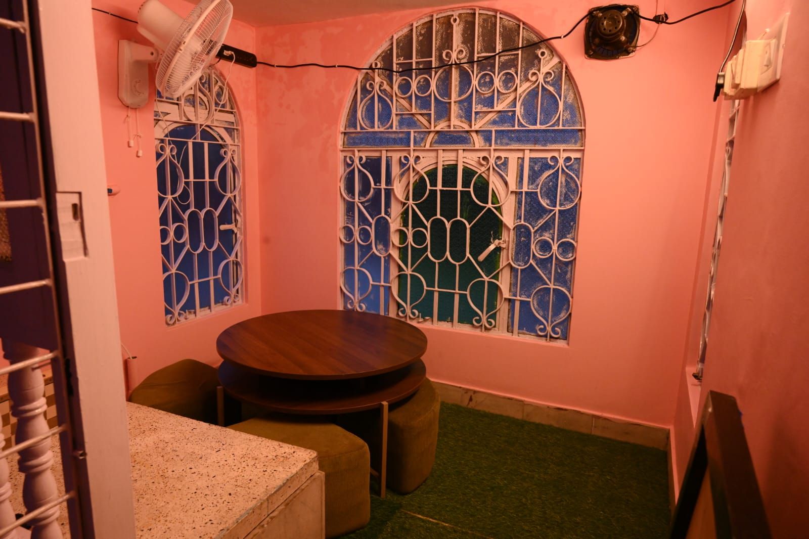 Ein kleiner Raum mit einem runden Tisch, zwei Stühlen, einem Ventilator und vergitterten Fenstern.