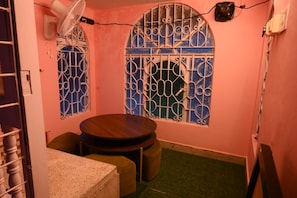 Una habitación pequeña con una mesa redonda, dos sillas, un ventilador y ventanas con barrotes.