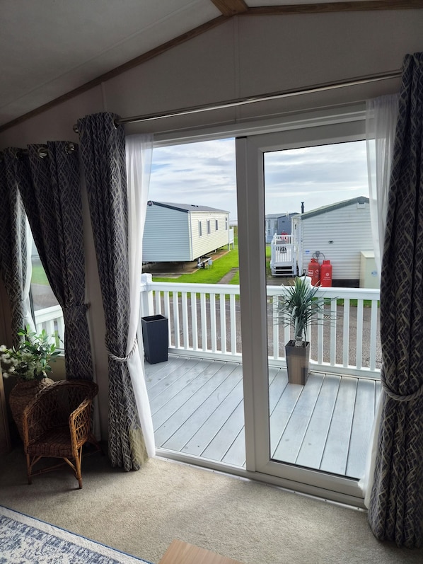 A room with a sliding glass door leading to an outdoor deck, a wicker chair, a potted plant, and a trash bin. - Luxury Sea View Lodge (Southerness)