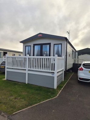 A caravan with a white deck and a grey exterior, parked on a grassy area with a paved path. - Luxury Sea View Lodge (Southerness)