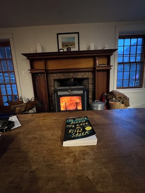 Una habitació amb una llar de foc, una taula amb un llibre anomenat 'The House Across the Lake' de Riley Sager, i una finestra amb vista a les muntanyes.