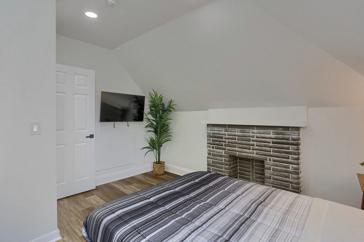 A bedroom with a bed, a fireplace, a potted plant, and a television on the wall.