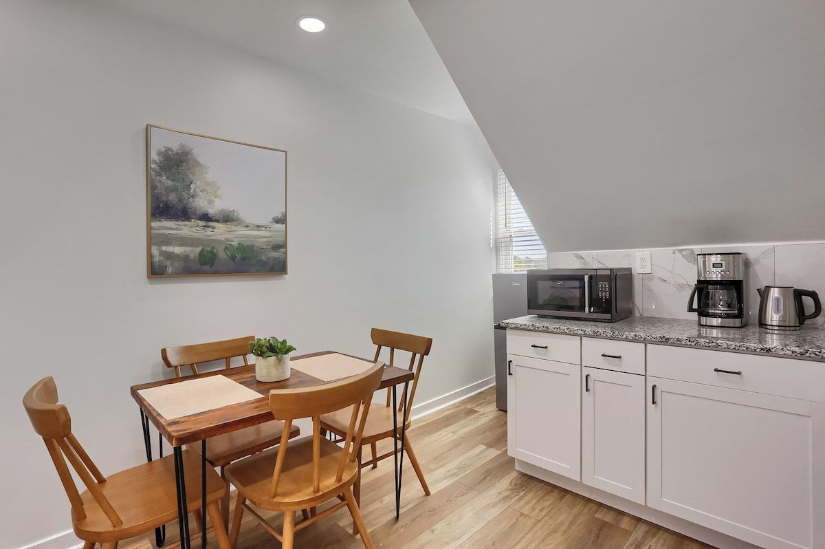 A modern kitchen with white cabinets, a microwave, a coffee maker, and a dining area with a wooden table and chairs.