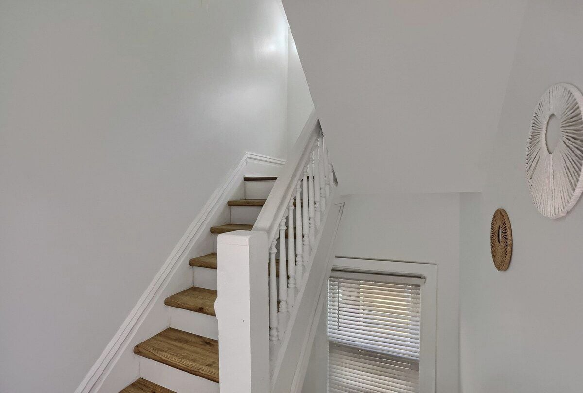 A staircase with wooden steps and white railings, leading to a room with a white ceiling and a window with blinds.