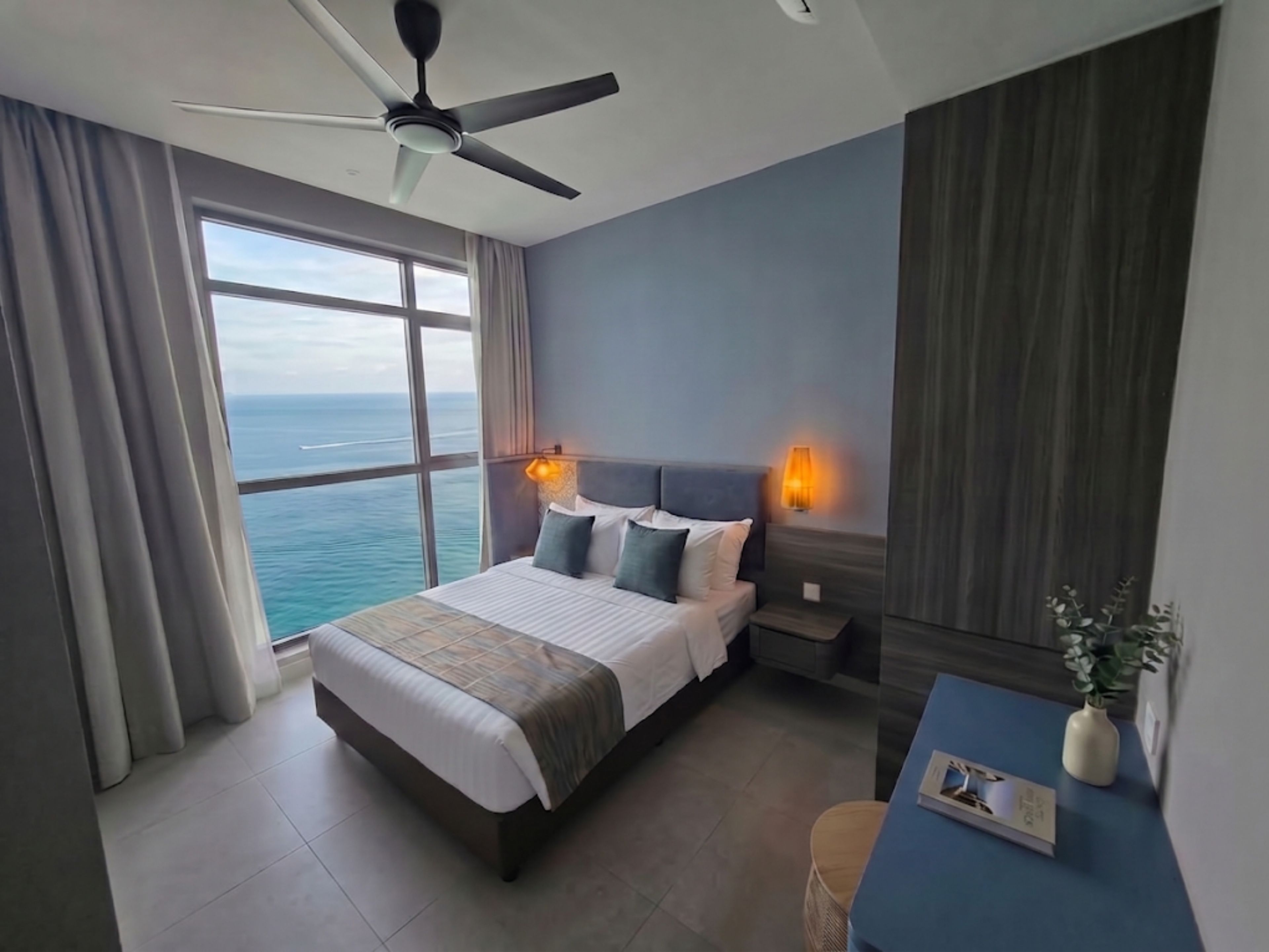 A hotel room with a bed, a desk, a chair, a ceiling fan, a nightstand, a lamp, and a view of the ocean.